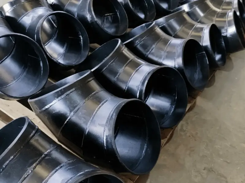 Lote de curvas tubulares e conexões de aço com revestimento de proteção preto de alto padrão, alinhadas em paletes na fábrica do Grupo SAG, prontas para montagem final.