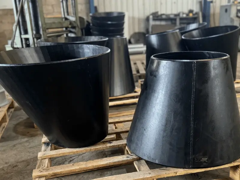 Reduções concêntricas e excêntricas de aço com revestimento industrial preto, organizadas sobre paletes de madeira no galpão de expedição do Grupo SAG.